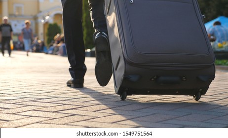 Feet Of Successful Businessman Walking Through City Street And Pulling Suitcase On Wheels At Sunny Day. Confident Man On Way To His Flight. Male Legs In Shoes Stepping At Urban Environment. Slow Mo