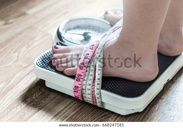 Feet tied up with measuring tape to a weight\
scale. Addiction and obsession to weight loss. Anorexia and eating\
disorder concept.