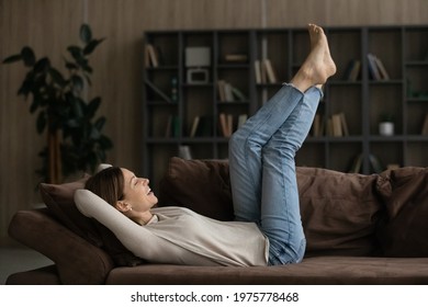 Feet up. Casual young female lie barefoot on soft comfy couch relax raise legs up do lazy gymnastic exercises to normalize blood circulation. Carefree millennial woman rest on sofa enjoy doing nothing