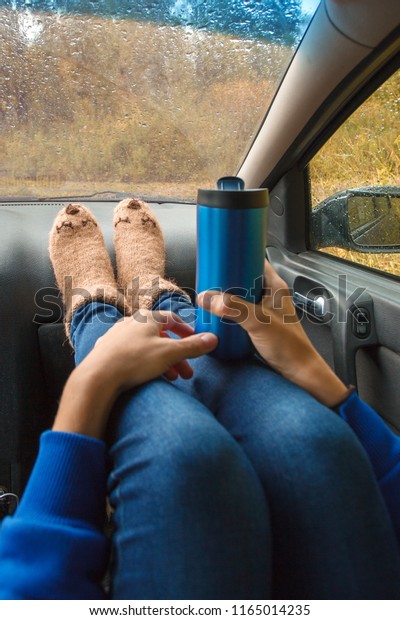 Feet in warm cute socks on car dashboard. Travel,\
road trip and autumn fall concept. Focus on thermos bottle cup with\
hot drink in female hands.\
