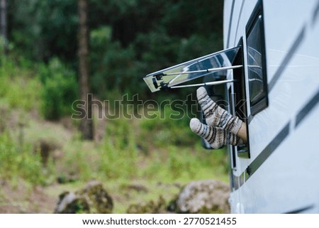 Feet in warm patterned socks extend from an open camper window while parked in a green forest, conveying cozy relaxation, outdoor holiday vibes and slow, peaceful moments on a camping adventure.