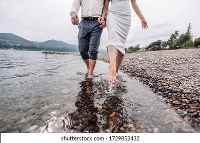 Feet In Water. Two People Walking With Naked Legs On River Shore. Pebbles Under Bare Foot. Cold Water Of River.