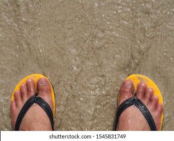 The feet wear yellow slippers, standing on the sandy beach and the sea water blows in.