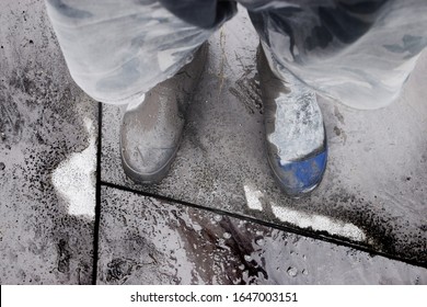 Feet Wearing Dirty Gumboots On A Worksite Surrounded By Concrete Dust And Dirt, With Deep Cuts In The Concrete Driveway