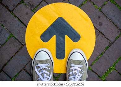 Feet Wearing Sneakers In Front Of Arrow On Road Outdoors. Direction Arrow In Front Of A Store For The Social Distancing During The Covid-19 Pandemic. One Way