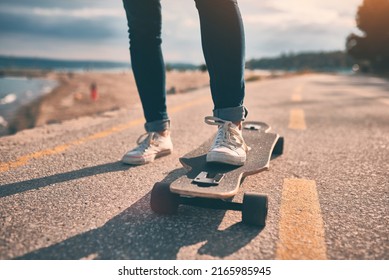 Feet of woman in sneakers on a double kick cutaway longboard in the city against the backdrop of the sea in the evening
