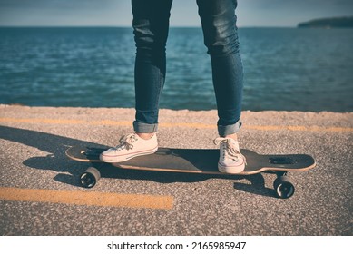 Feet of woman in sneakers on a double kick cutaway longboard in the city against the backdrop of the sea in the evening