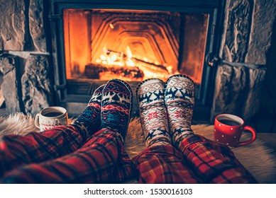 Feet in woollen socks by the Christmas fireplace. Family sitting relaxes by cozy authentic fireside with a cup of hot drink and warming up their feet. Winter and Christmas holidays concept - Powered by Shutterstock