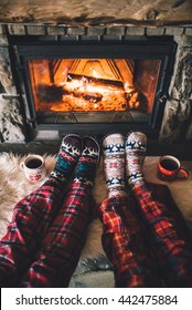 Feet In Woollen Socks By The Christmas Fireplace. Family Sitting Relaxes By Cozy Authentic Fireside With A Cup Of Hot Drink And Warming Up Their Feet. Winter And Christmas Holidays Concept