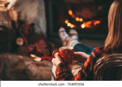 Feet in woollen socks by the Christmas fireplace. Woman relaxes by warm fire with a cup of hot drink and warming up her feet in woollen socks. Close up. Winter and Christmas holidays concept.
