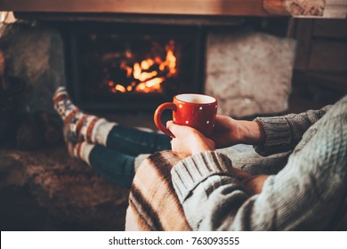 Feet in woollen socks by the Christmas fireplace. Woman relaxes by warm fire with a cup of hot drink and warming up her feet in woollen socks. Cozy atmosphere. Winter and Christmas holidays concept.