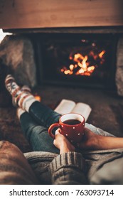 Feet In Woollen Socks By The Christmas Fireplace. Woman Relaxes By Warm Fire With A Cup Of Hot Drink And Warming Up Her Feet In Woollen Socks. Cozy Atmosphere. Winter And Christmas Holidays Concept.