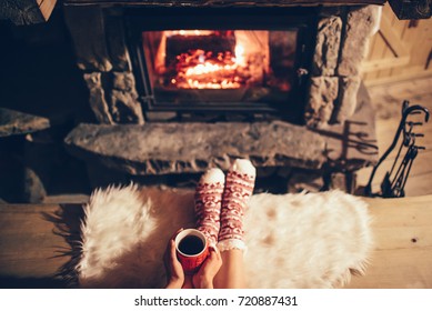 Feet In Woollen Socks By The Cozy Christmas Fireplace. Woman Relaxes By Warm Fire With A Cup Of Hot Drink And Warming Up Her Feet In Woollen Socks. Winter And Christmas Holidays Concept.