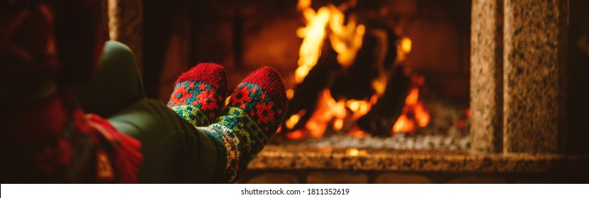 Feet In Woollen Socks By The Fireplace. Woman Relaxes By Warm Fire And Warming Up Her Feet In Woollen Socks. Staying At Home During Quarantine. Winter And Christmas Holidays Concept. Banner Format.