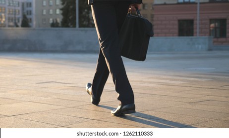 Feet Of Young Businessman With A Briefcase Walking In City Street. Business Man Commuting To Work. Confident Guy In Suit Being On His Way To Work. Cityscape Backround. Slow Motion Rear View Close Up.