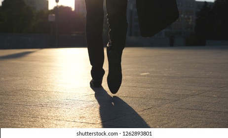 Feet Of Young Businessman With A Briefcase Walking In City Street. Business Man Commuting To Work. Confident Guy In Suit Being On His Way To Work. Cityscape Backround. Slow Motion Rear View Close Up.