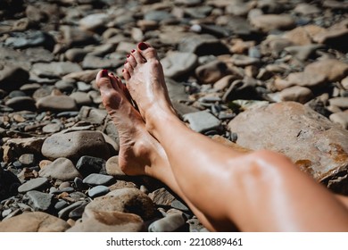 The Feet Of A Young Girl Lie On A Stone Beach, A Man Sunbathes Under The Sun, The Lower Part Of The Human Body, Women's Feet, A Red Pedicure. High Quality Photo