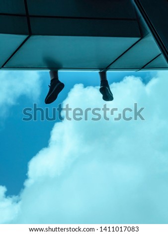 Feet of young man who sits on ledge of skyscraper, facing clouds and blue sky, for urban, travel, or psychological concepts (selective focus)