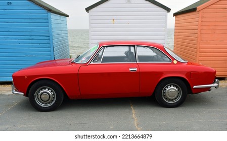 FELIXSTOWE, SUFFOLK, ENGLAND -  MAY 01, 2022: Classic Red Alfa Romeo Motor Car Parked In Front Of Beach Hut On Seafront Promenade