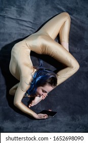 A Female Acrobat Lies On A Gray Bedspread In A Curved Pose And Uses A Mobile. A Woman Contortionist Resting On A Bed In An Acrobatic Stunt And Texting On The Phone.