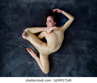 A Female Acrobat Lies On A Gray Bedspread In A Curved Pose And Uses A Mobile. A Woman Contortionist Resting On A Bed In An Acrobatic Stunt And Texting On The Phone.