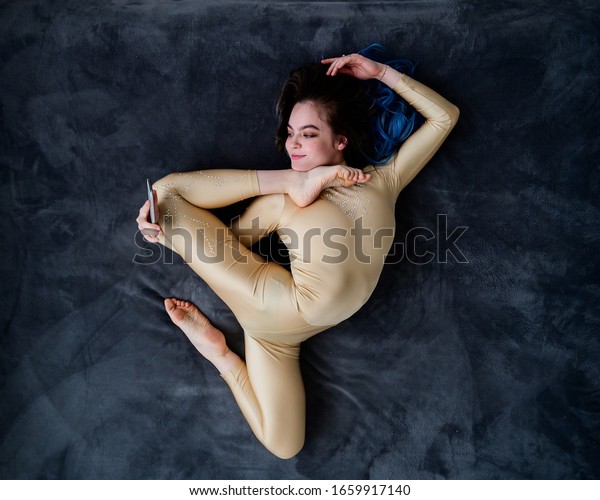 A female acrobat lies on\
a gray bedspread in a curved pose and uses a mobile. A woman\
contortionist resting on a bed in an acrobatic stunt and texting on\
the phone.