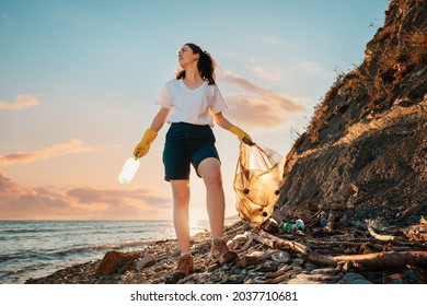 A female activist stands with a plastic bag and a bottle in her hand against the background of the sunset. Bottom view. Cleaning of the coastal zone and wild beaches. The concept of Earth Day.