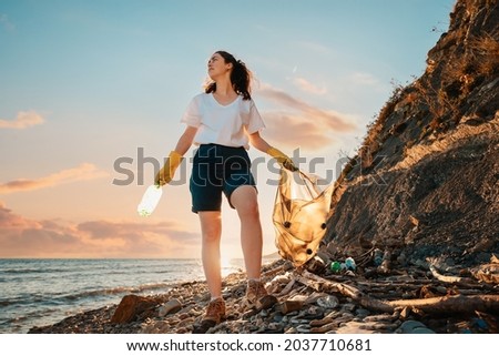 A female activist stands with a plastic bag and a bottle in her hand against the background of the sunset. Bottom view. Cleaning of the coastal zone and wild beaches. The concept of Earth Day.
