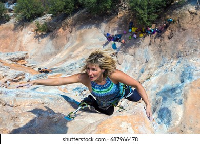 Female Adult Climber In Blue Shirt And Black Pants Reaching Next Hold On High Vertical Rock Using Safety Gear And Climbing Shoes