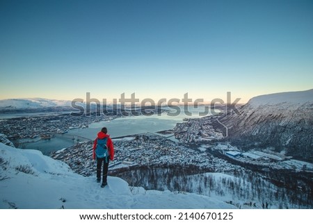Female Adventurer in a red jacket and backpack stands atop a fjellheisen and watches Norwegian Sea surrounding the city of Tromso in northern Norway above the Arctic Circle on frosty winter morning.