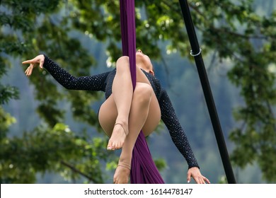 A Female Aerial Contortionist Is Seen Suspended From Silk Fabric During A Performance In Nature, With Blurry Trees In Background And Copy Space To Right