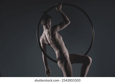 A female aerial hoop artist demonstrates poise and strength, gracefully balancing in the hoop. Her toned physique, tattoo, and expressive pose are highlighted against a dark studio backdrop - Powered by Shutterstock