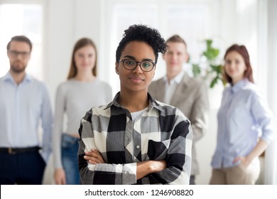 Female African American millennial employee standing foreground with arms crossed, colleagues at background, black smiling young woman professional look at camera, successful woman posing in office