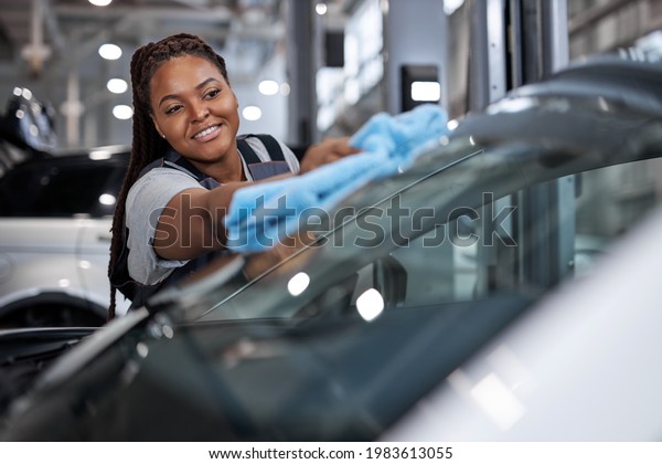 Female african auto mechanic\
cleaner is cleaning car window glass with blue rag, side view on\
adorable black woman in car service, working alone, washing\
vehicle