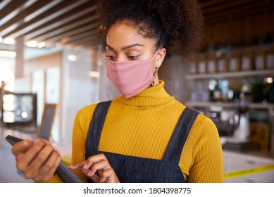 Female African coffee shop owner wearing face mask standing in front of counter performing stock check.