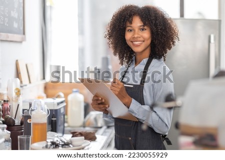 Female African coffee shop small business owner wearing apron standing in front of counter performing stock check. afro hair employee Barista entrepreneur.