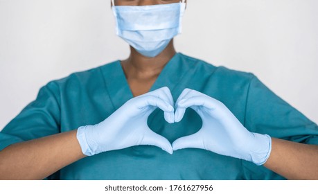 Female african professional medic nurse wear face mask, gloves, blue green uniform showing heart hands shape. Medical love, care and safety symbol, corona virus health protection sign concept. Closeup