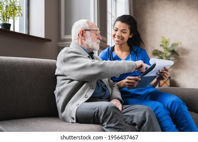 Female african-american professional doctor showing medical test result explaining prescription using clipboard visiting senior elderly old man patient at home sitting on sofa. 