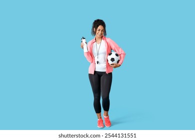 Female African-American soccer trainer with ball and water bottle on blue background - Powered by Shutterstock