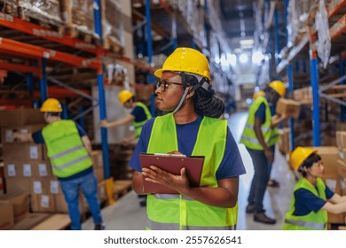 Female African-American warehouse worker doing inventory, holding a clipboard, wearing a safety vest and a hardhat while other workers are busy behind her. - Powered by Shutterstock