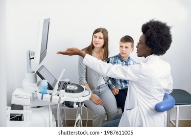 Female Afro American Ultrasound Doctor Pointing At Monitor Of Usg Machine And Explaining Results Of Regular Check Up For Her Patients, Caucasian Woman And Her Teen Son