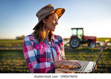 Female agronomist using laptop modern technology in agriculture. Young farmer standing in field in front of tractor machinery smiling.  - Powered by Shutterstock