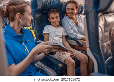 Female Air Hostess Trying To Entertain A Kid On The Plane By Offering A Book To Read. Cabin Crew Provide Service To Family In Airplane. Airline Transportation And Tourism Concept