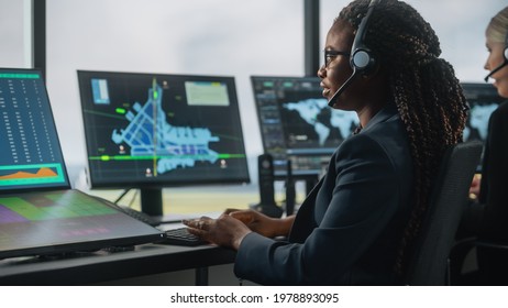 Female Air Traffic Controller With Headset Talk On A Call In Airport Tower. Office Room Is Full Of Desktop Computer Displays With Navigation Screens, Airplane Departure And Arrival Data For The Team.