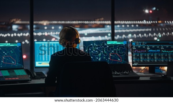 Female Air Traffic Controller with Headset Talk on\
a Call in Airport Tower at Night. Office Room is Full of Desktop\
Computer Displays with Navigation Screens, Airplane Flight Radar\
Data for the Team.