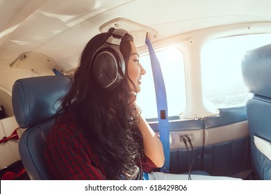 Female Airplane Passenger Enjoying The View From The Cabin Window Of Small Sport Plane. Young Brunette Woman In Headphones Sitting In Plane During Flight