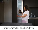 Female with alcohol addiction drinking alcohol in the morning. Alcoholism, mental health and social problems. Woman suffers from dipsomania, taking out a bottle of wine from the fridge at home kitchen