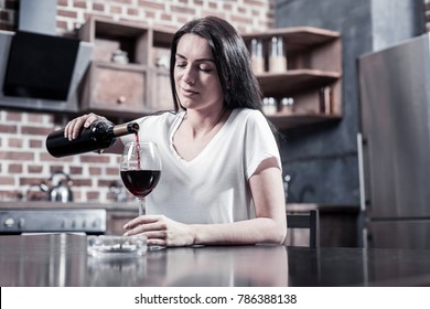 Female alcoholism. Sad cheerless brunette woman sitting at the table and holding a bottle while pouring wine