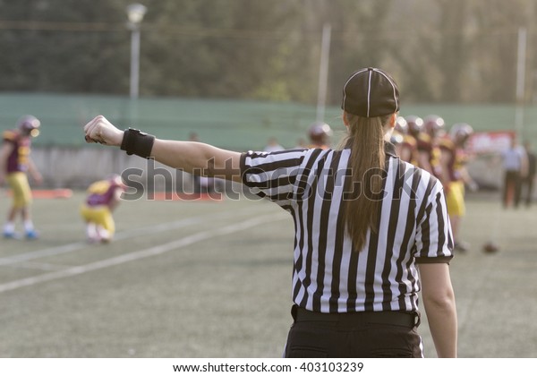 Female American football referee giving\
signals and blurred players in the\
background
