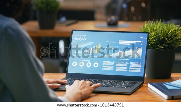 Female Analyst at Her Desk Works on\
a Laptop Showing Statistics, Graphs and Charts. She Works on the\
Wooden Table in Creative Office. Over the Shoulder\
Footage.